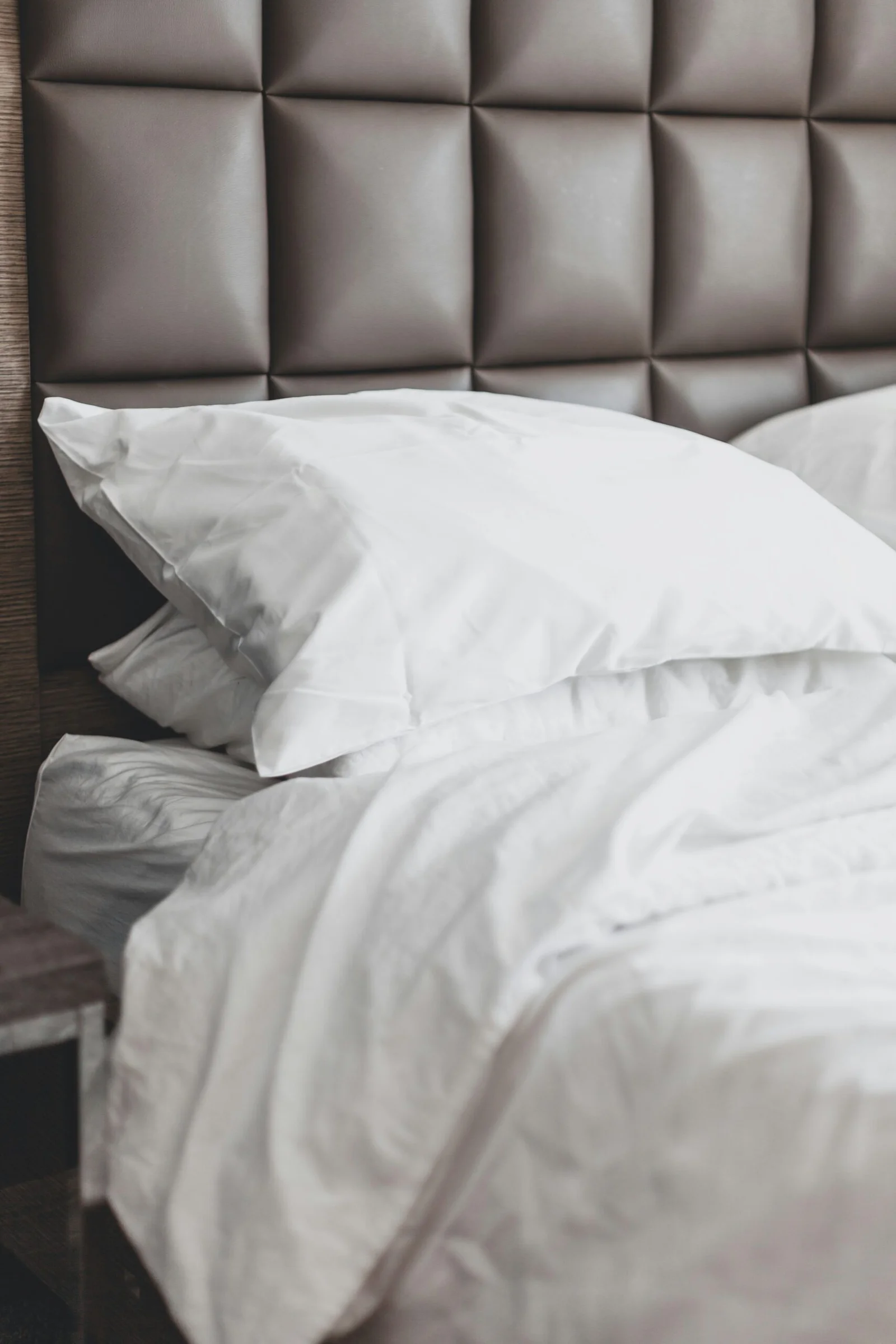 A cozy bedroom scene featuring plush white Beckham Hotel Collection pillows on a bed with a grey tufted leather headboard.